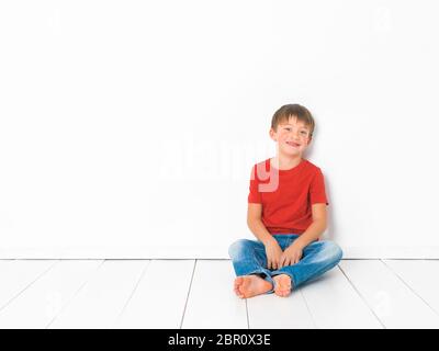 Et mignon garçon blond avec chemise rouge et bleu jeans pose sur plancher en bois blanc in front of white background Banque D'Images