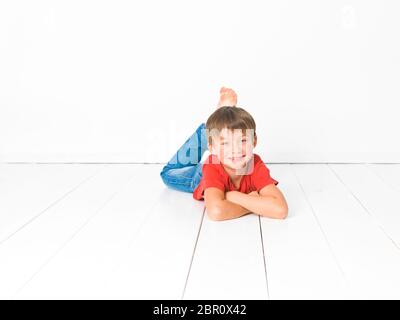 Et mignon garçon blond avec chemise rouge et bleu jeans pose sur plancher en bois blanc in front of white background Banque D'Images