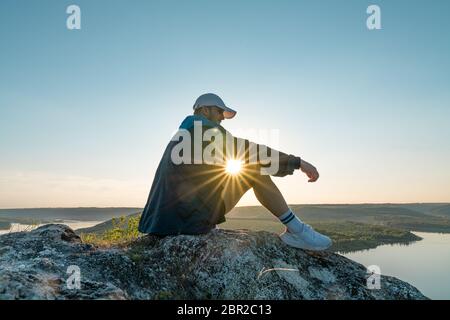 Homme assis sur une perge de montagne, profitant du beau coucher de soleil sur une large vallée de rivière Banque D'Images