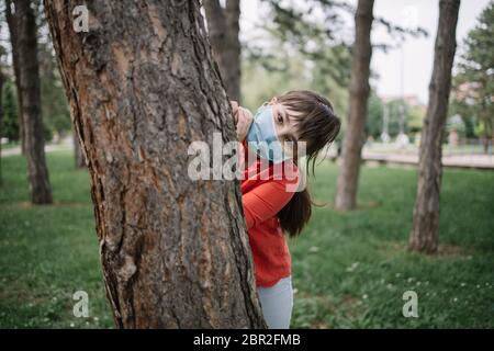 Fille portant un masque antivirus se cachant derrière l'arbre. Portrait d'un enfant avec masque médical jouant dans le parc pendant une pandémie de coronavirus. Banque D'Images