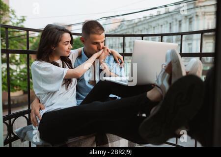 Assis sur le balcon, enserrer, regarder des séries. Quarantaine verrouillage, séjour à la maison concept - beau couple caucasien appréciant un nouveau style de vie pendant le coronavirus. Bonheur, ensemble, santé. Banque D'Images