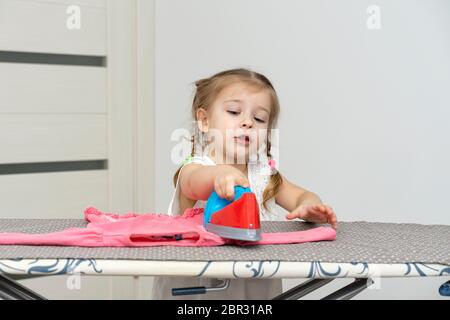 une jolie petite fille de 4 ans joue une femme au foyer, repassant les vêtements des enfants avec un fer à repasser Banque D'Images