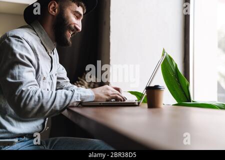 Photo de rire jeune homme portant un chapeau tapant sur l'ordinateur portable et boire du café tout en étant assis à la table dans le café à l'intérieur Banque D'Images