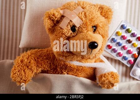 Petit ours blessé gisant malade au lit avec un plâtre sur son front et bandée bras en écharpe à côté d'une plaquette de pilules de couleur Banque D'Images