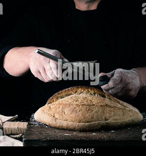 Chef en uniforme noir aiguise un couteau de cuisine, à côté d'elle est cuite la farine de blé blanche, fond noir Banque D'Images