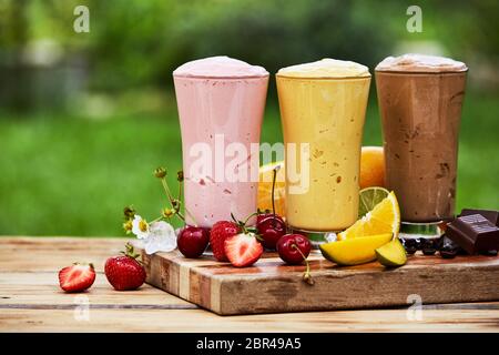 Trois délicieux fruits tropicaux frais et de chocolat milk-shakes et smoothies servi dans des grands verres à l'extérieur sur une planche en bois dans le jardin Banque D'Images