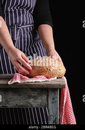 Femme dans un tablier à rayures bleu tient dans ses mains le pain de seigle rond de boulangerie et de graines de tournesol, table en bois Banque D'Images