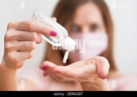 Jeune femme avec du coton rose fait maison bouche visage virus masque compte-gouttes anti bactérien alcool gel à frotter sur la main, détail à la bouteille blanche, visage flou Banque D'Images