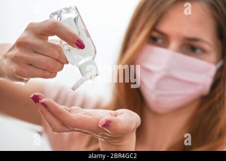 Jeune femme avec du coton rose fait maison bouche visage virus masque compte-gouttes anti bactérien alcool gel à frotter sur la main, détail à la bouteille blanche, visage flou Banque D'Images