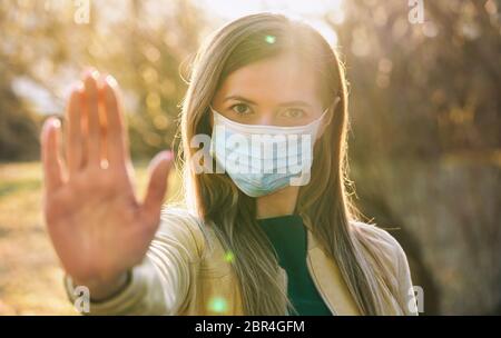 Jeune femme portant un masque de nez bleu jetable à usage unique, porte la main devant elle comme geste d'arrêt, coucher de soleil flou allumé les arbres en arrière-plan. Banque D'Images