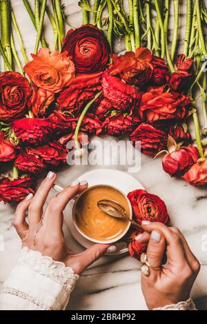 Plat de femme tenant main tasse de café expresso noir et cuillère sur table en marbre blanc avec tas de fleurs de ranunculus rouges, vue du dessus. Saisonnier Banque D'Images