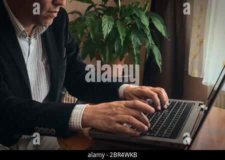 Gros plan sur un homme adulte senior vêque de vêtements élégants qui travaillent à la maison en utilisant son ordinateur portable sur une table pendant qu'il est assis sur un fauteuil avec une plante Banque D'Images