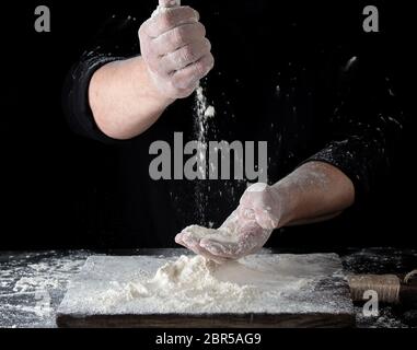 Chef en uniforme noir examine ses doigts la farine de blé blanche au-dessus de la table, fond noir Banque D'Images