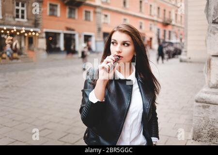 Une fille forte dans une veste en cuir noir fume une cigarette électronique dans une rue de ville Banque D'Images