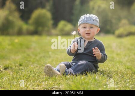 Un bébé caucasien aux yeux bleus en chapeau est assis sur une route de prairie et donne une Marguerite à la caméra. Banque D'Images