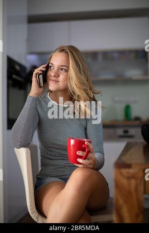 Adolescente appelant sur son téléphone portable tout en ayant une tasse de thé dans une cuisine moderne (shalllow DOF, tons de couleur libre) Banque D'Images