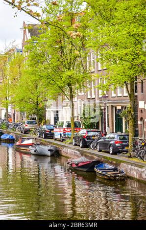 Amsterdam, pays-Bas - 27 avril 2019 : petit bateau sur le canal dans le centre-ville de la capitale néerlandaise. Voitures garées par des arbres verts le long du canal. Photo verticale. Banque D'Images