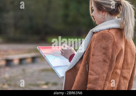 Une jeune fille dessine une image directement dans le parc. Peintre femme dans une veste brune se tient dans un parc tenant une peinture dans les mains la dessinant Banque D'Images