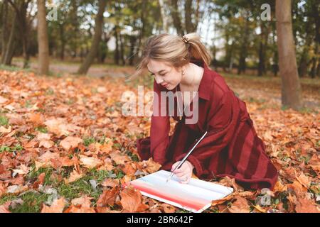 Jeune artiste dessine une image agenouillée dans un parc sur l'herbe. Fille s'inspire de la créativité assis dans le parc d'automne et de la peinture Banque D'Images