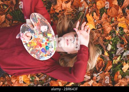 Le peintre se trouve sur l'herbe dans le parc d'automne, tenant une brosse et une palette dans sa main. Un jeune artiste se détend dans un parc sur l'herbe Banque D'Images