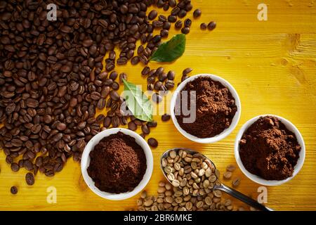 Les grains de café torréfié et de matières premières avec de nouveaux motifs dans des petits bols vue d'en haut sur bois jaune avec deux feuilles vertes et copy space Banque D'Images