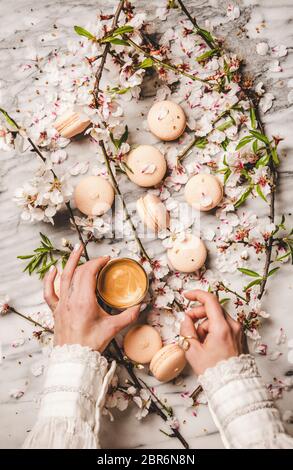 Une coulées de femmes tient une tasse de café et des biscuits au macaron sur des fleurs de printemps blanches et un fond de marbre blanc, vue du dessus Banque D'Images
