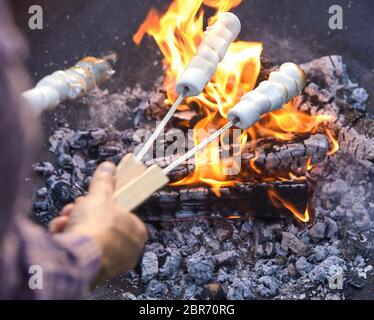 Man toasting marshmallows grésillant sur les brochettes en métal sur la braise d'un barbecue ou feu de camp en vue de l'épaule de son passé de bonbons Banque D'Images