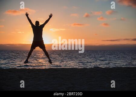 Une vue arrière-plan d'un homme méconnaissable faisant sauts sur la plage, il est à l'extérieur, vers la mer et le coucher du soleil sur une belle soirée dans Banque D'Images