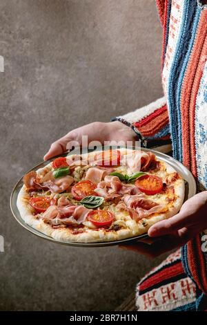 Pizza maison fraîche avec jambon prosciutto, fromage, tomates, basilic sur plaque dans les mains des femmes. Cuisson à domicile ou livraison de fast-food. Banque D'Images