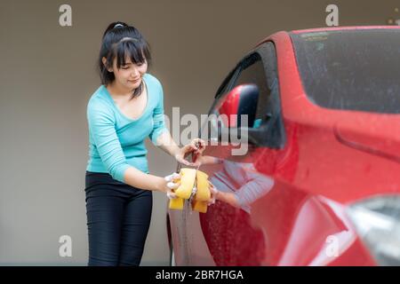 Une femme asiatique ayant du plaisir à la maison laver la voiture rouge avec des suds et une éponge pendant son séjour à la maison en utilisant le temps libre sur leur entretien quotidien Banque D'Images