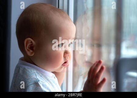 un petit garçon sur le rebord de la fenêtre regarde la fenêtre.un enfant avec une expression triste sur son visage Banque D'Images