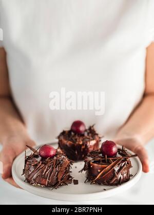 Femme en t-shirt blanc tient une assiette avec trois mini-gâteaux de forêt noire avec des éclats de cerise et de chocolat. Verticale. Copier l'espace haut Banque D'Images