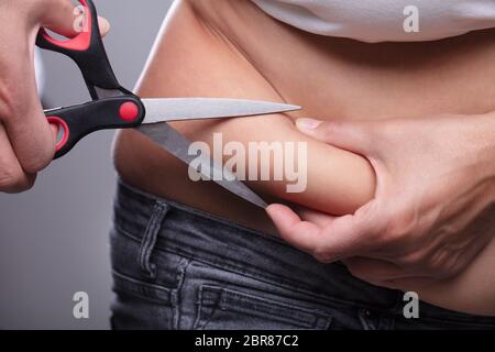Mid Section d'une femme de couper le trop de graisse de ventre avec Scissor contre fond gris Banque D'Images