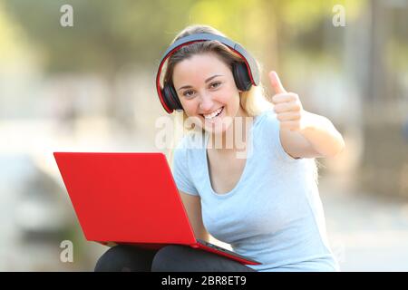 Happy girl with laptop and headphones with Thumbs up assis à l'extérieur dans un parc Banque D'Images