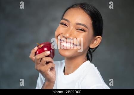 Portrait d'une jeune femme souriante en t-shirt blanc tenant une pomme rouge sur un fond en béton Banque D'Images