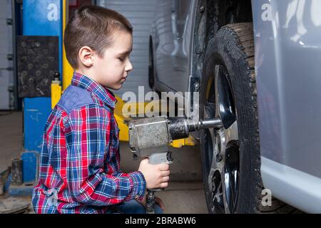 Un jeune garçon, un jeune travailleur automobile, fait un changement de pneu avec une clé pneumatique dans le garage d'une station de service. Un enfant apprend la changine mécanique Banque D'Images