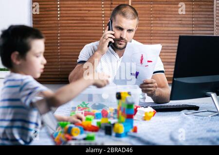 Un jeune papa parle au téléphone et essaie de travailler à un ordinateur à distance pendant la période d'auto-isolation en relation avec le coro Banque D'Images