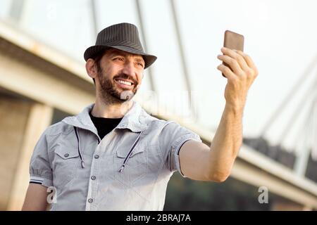 Homme moyen souriant décontracté en chapeau fait selfie sur lui avec téléphone mobile à l'extérieur. Banque D'Images