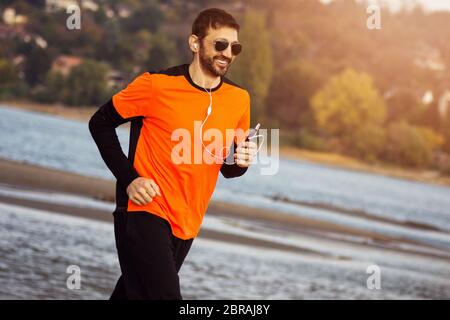Le sourire dans les vêtements de sport orange avec des écouteurs et des lunettes de soleil est le jogging au bord de la rivière, concept de loisirs. Banque D'Images