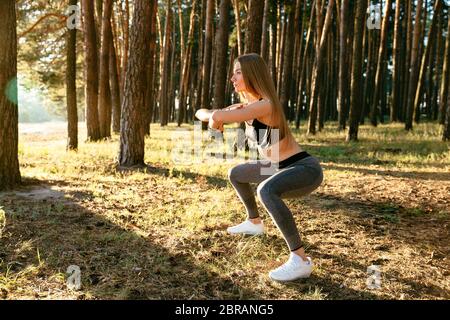 Vue latérale du sportif attrayant femme en haut du réservoir et des jambières, faire des squats, l'étirement du corps, de l'exercice dans le bois. Sport concept. Banque D'Images