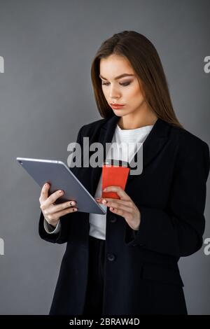 Une jeune fille en vêtements de style professionnel se tient sur un fond gris avec une tablette et une tasse de café dans ses mains Banque D'Images