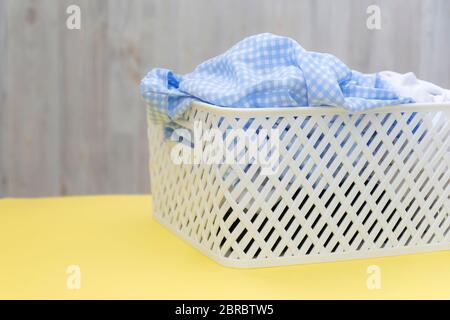 Chemises blanches bleues vierges dans un panier en plastique blanc sur une table jaune sur fond gris. Blanchisserie, nettoyage à la maison, propreté et fraîcheur Banque D'Images