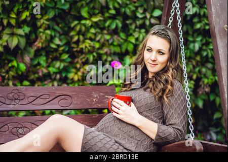 Une jeune femme enceinte assise sur une balançoire, avec une tasse de thé. Femme enceinte se détendant dans le parc. Banque D'Images