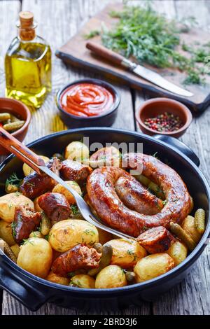 Saucisse de porc spirale rôtie au four avec pommes de terre et cornichons neufs, arrosée d'aneth frais sur un plat noir sur fond de bois avec ketchup, pepperco Banque D'Images