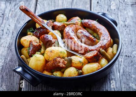 Saucisse italienne rôtie avec de nouvelles pommes de terre et des cornichons, arrosée d'aneth frais sur un plat de cuisson noir sur une table en bois, en gros plan Banque D'Images