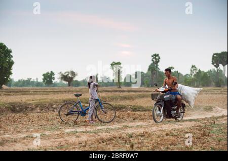 Une jeune fille cambodgienne s'arrête sur un vélo pour permettre à un homme cambodgien transportant des marchandises de passer sur un cyclomoteur sur une petite piste. Province de Siem Reap, Cambodge. Banque D'Images