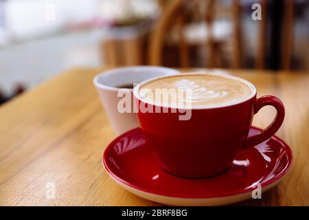 Café aromatique dans une tasse rouge avec mousse de lait et art latte et café fraîchement préparé dans une tasse blanche sur une table en bois clair. Banque D'Images