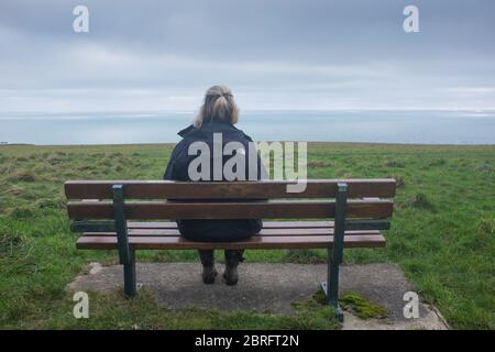 Une femme seule est assise sur un banc de parc près de la côte et donne sur la mer au loin Banque D'Images