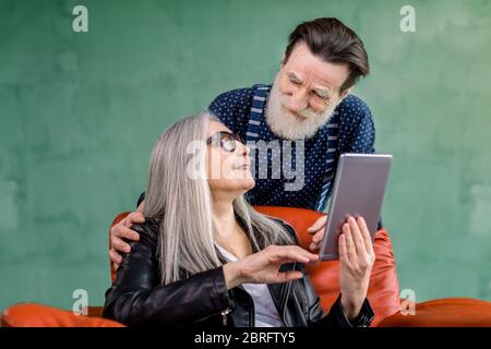 Photo en studio d'un couple de personnes âgées heureux, homme et femme stylés, regardant les uns les autres tout en naviguant sur Internet ou en utilisant des applications sur une tablette i-pad et en parlant Banque D'Images