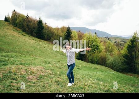 Heureuse fille touristique souriante dans les montagnes entourées de forêt, levant les armes et appréciant le silence et l'harmonie de la nature Banque D'Images